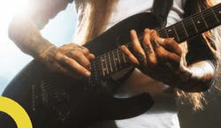 Imagem de homem com as mãos e braços tatuados e cabelos castanhos compridos usando uma camiseta branca enquanto toca uma guitarra preta.