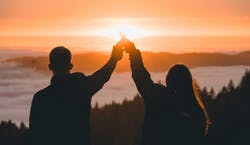 Imagem da sombra de um homem e uma mulher juntando suas mãos no ar enquanto observam o por do sol, que possui tons alarajados. 