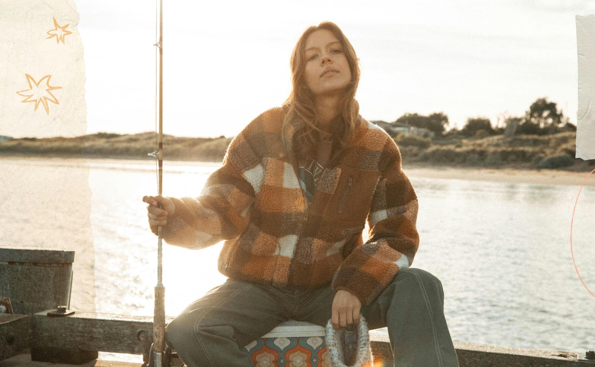 Image of female model sitting on an esky at the end of a pier holding a vintage fishing rod. She is wearing the Vacay Crew under the Outback Jacket with the Commando Pants.