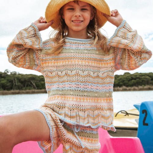 Young girl stands on bright pink paddle boat wearing the matching Annika Knit Top and Shorts in Dreamer Stripe. She holds a yellow hat on her head. 