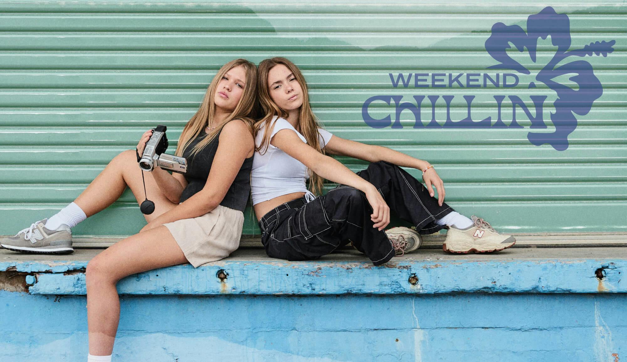 Image of two teen girls sitting on blue wall with a green roller door behind them. They lean back onto each other. The girl on the left wears a black singlet and natural linen shorts, the girl on the right wears a white crop tee and black cargo pants. graphic text sits in the top right reading 'weekend chillin'