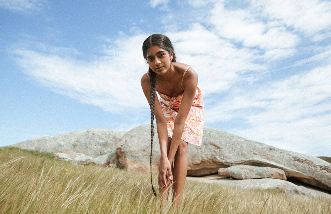 Female model wearing the Mia Mini dress in Pink Lizzie Paisley, leaning down towards the camera hands are crossed, with rocky country side in the background