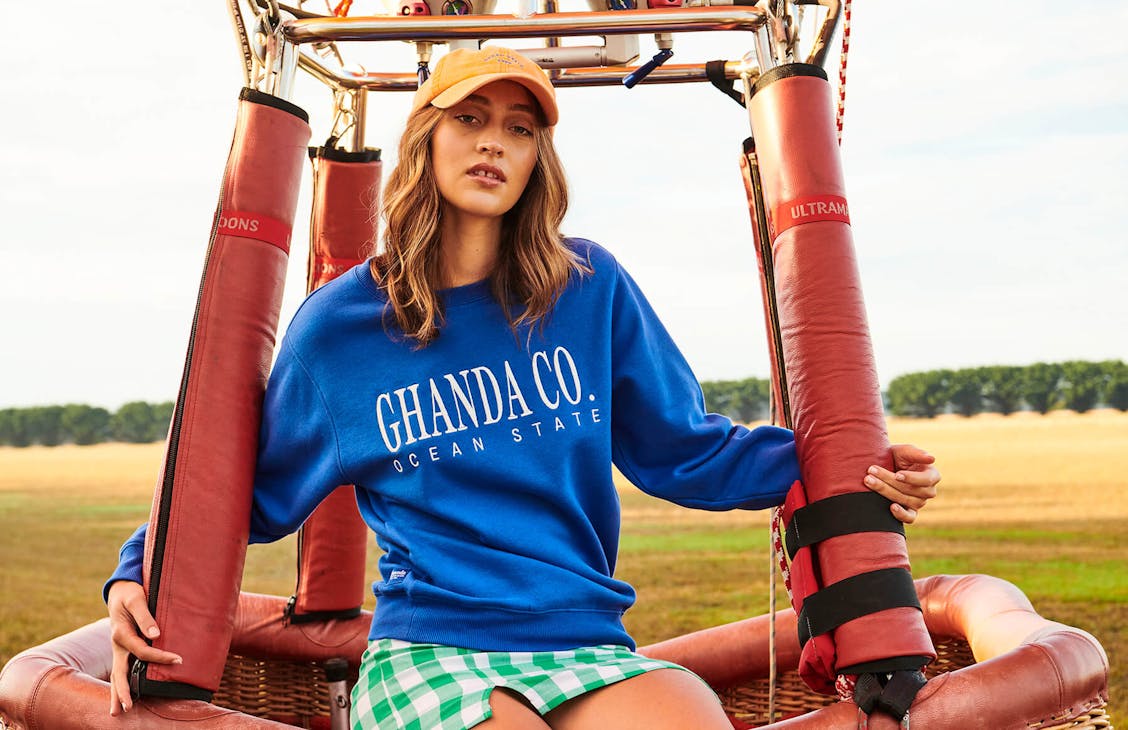 Image of model sitting on the outside of a hot air balloon basket. She is wearing the school skirt in t-rex green check, the classic cap in cord orange and the classic crew in pigment blue.