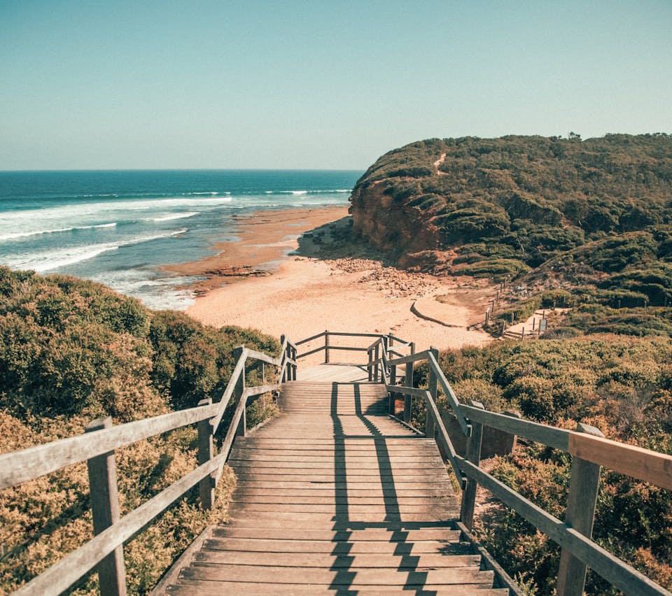 Photograph from top of stairs, leading down to beach. Blue ocean and grassy hills surround wooden steps.