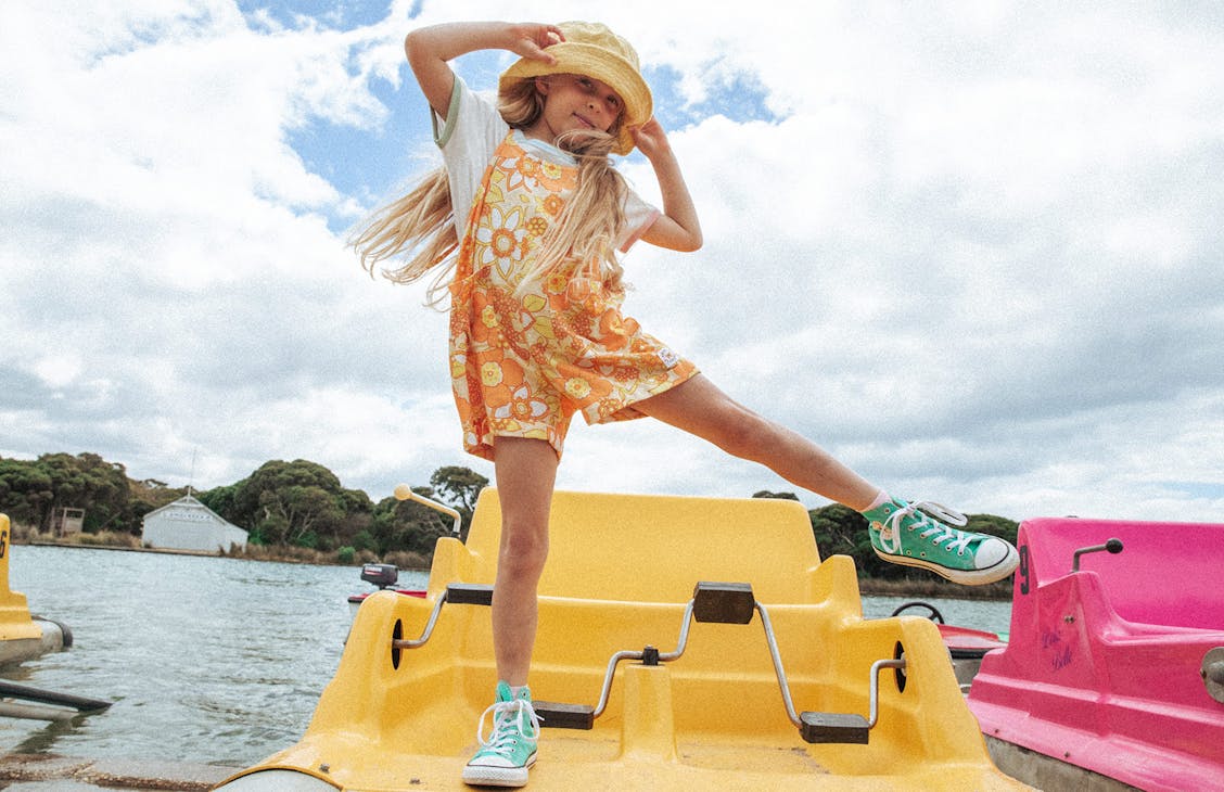 Young girl stands on a yellow paddle boat swinging her legs out. She wears the Circus Playsiut and the Terry Bucket Hat
