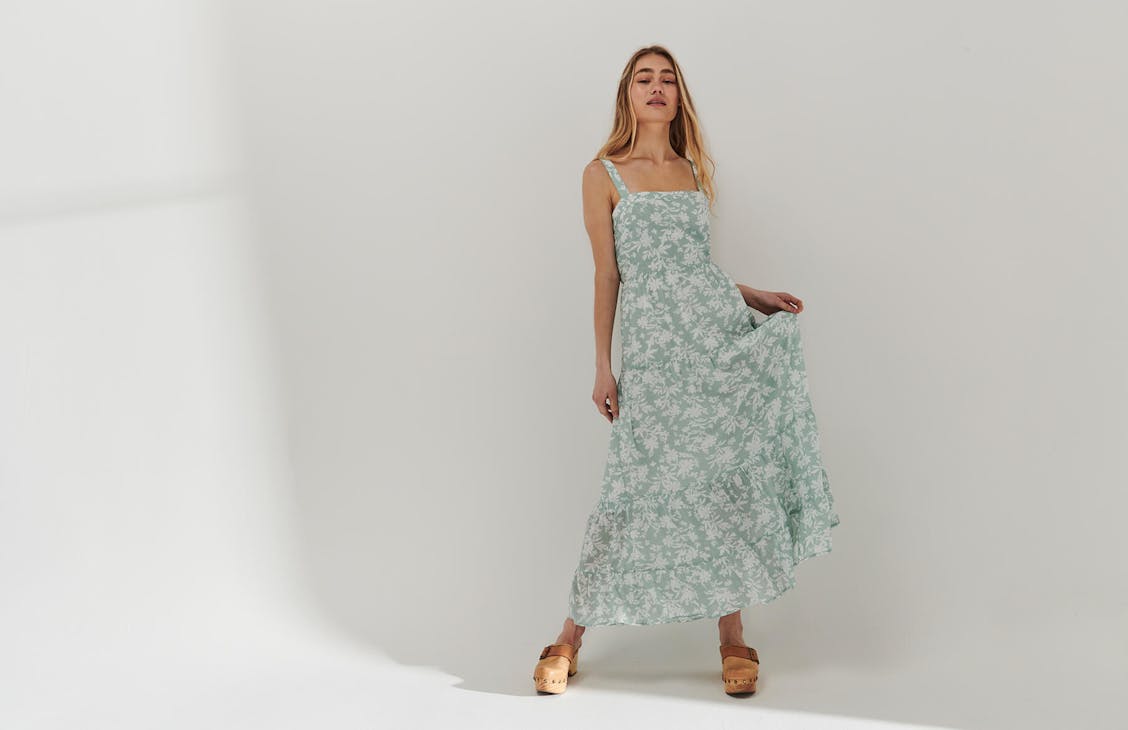 Female model wearing the Luella Dress in Wattle Floral eggshell, holding the dress up softly with her left hand.