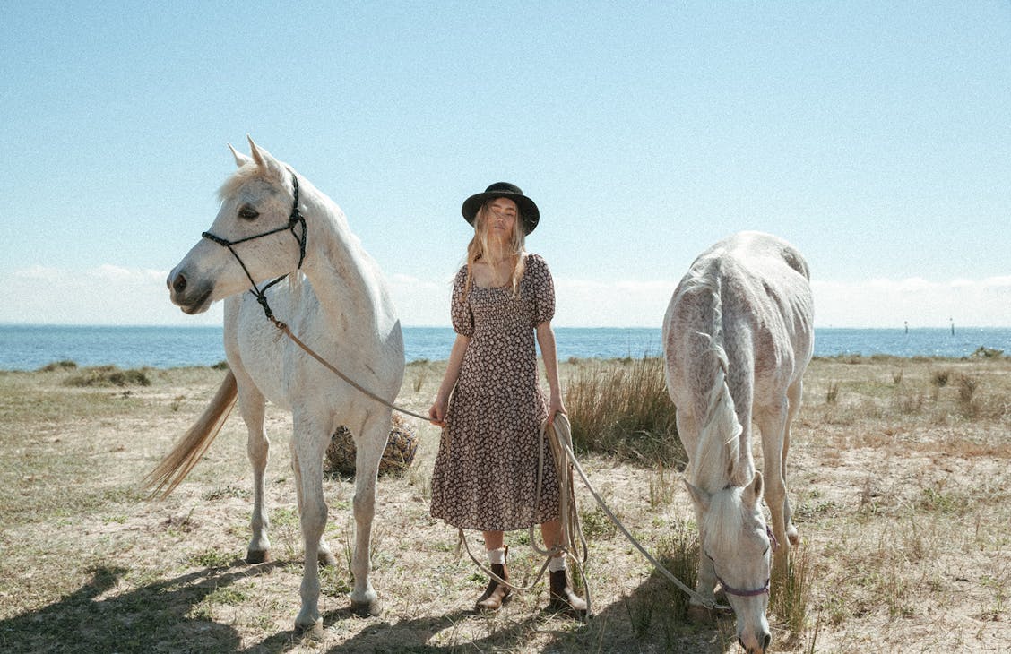 Female Model wearing the Eve Midi Dress in Brown Floral, standing between two white horses holding onto their rope neck ties, beach background behind