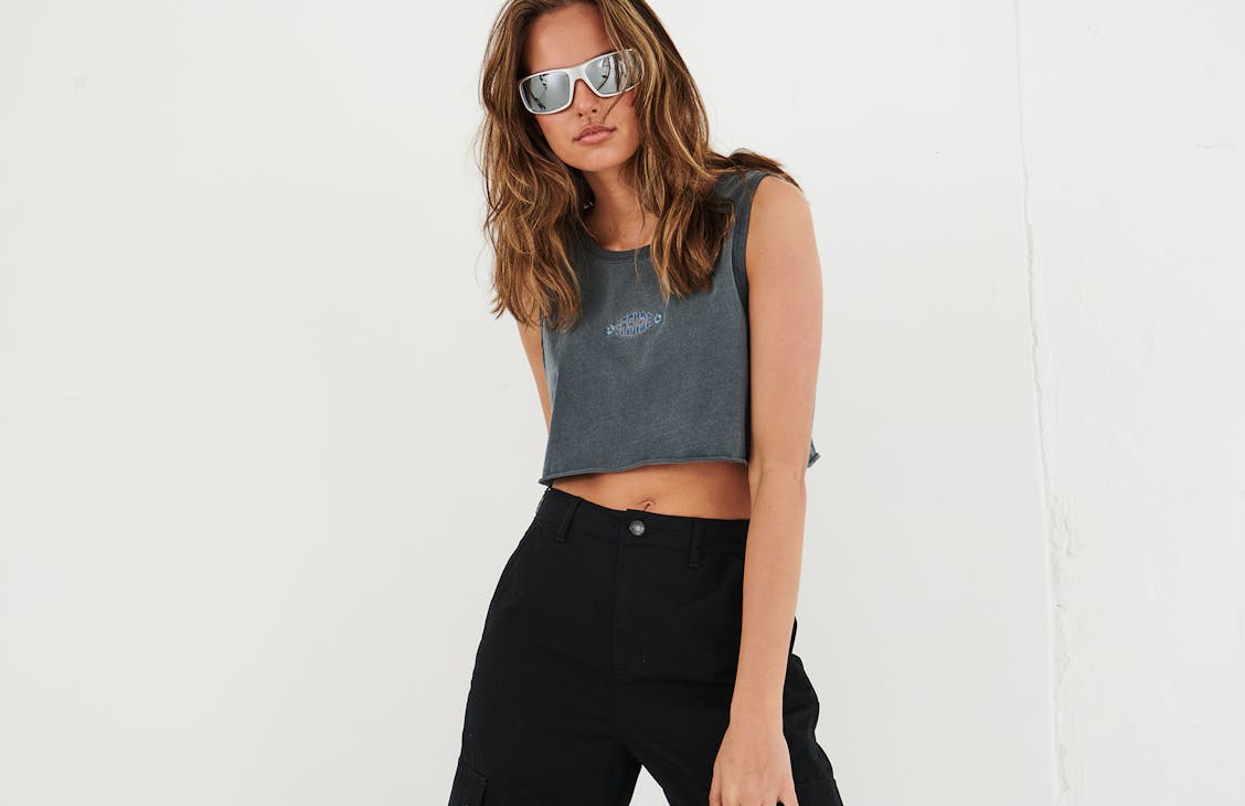 model in studio wearing a cropped grey singlet, with black pants and sunglasses.