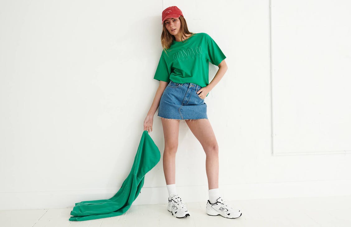 Image of female model in studio wearing a blue denim skirt and a green tshirt, she holds a green hoodie in her hand and wears a red cap