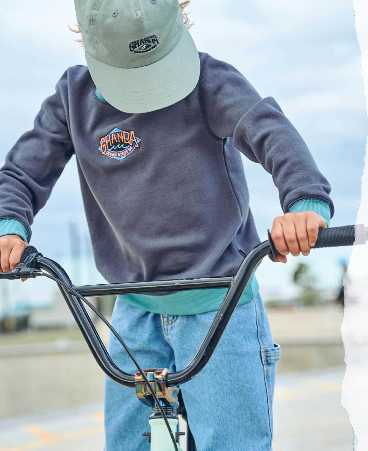 Image of young boy at the skate park on a bike. He is wearing denim jeans a ghanda embroidered panel crew and washed jade coloured cap