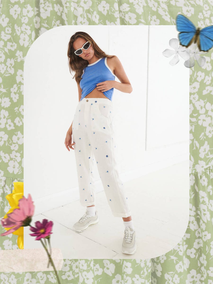 Image of model wearing white linen pants with small blue flower embroideries and a blue ringer singlet. The image if in an arched frame and in the background is a green and white floral. Other collage style flowers are around the frame