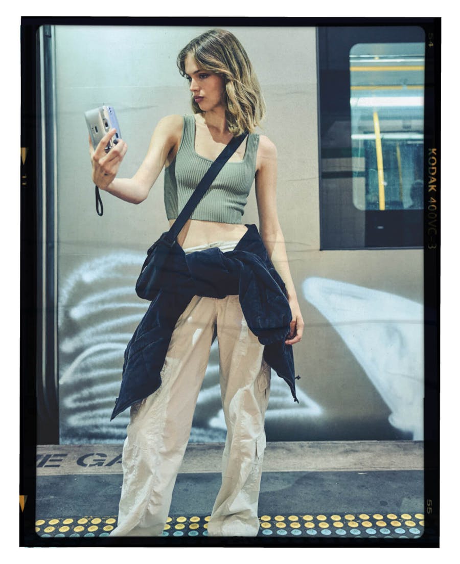 Image of female model taking selfie in the train station. She is wearing cargo pants a cropped green cami and a black cord jacket tied around her waist . Image is in a black frame