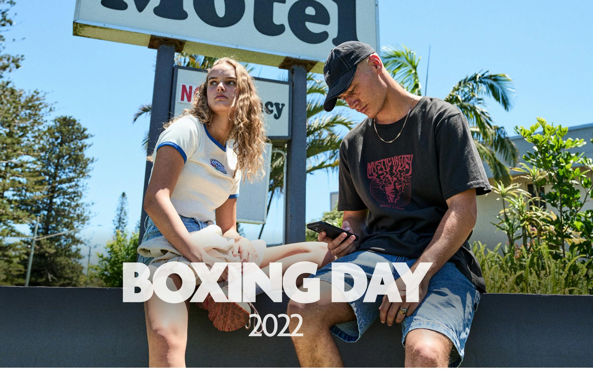 Image of a male and female model sitting on a ledge in front of a sign that reads 'Motel'. The text overlay on the image reads "Boxing Day 2022". 
The female is wearing our Crazy Crop Tee and denim shorts. The male is wearing our Mist Vintage Tee, Dogtown Shorts and Contrast Cap.