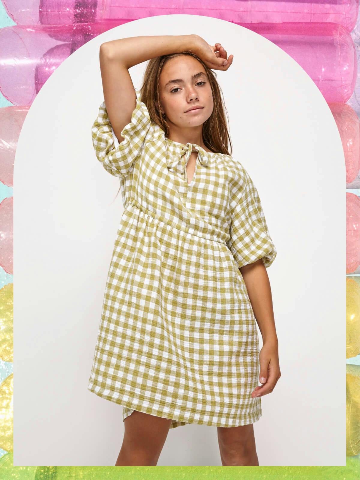 Image of teen model wearing a green check dress with puff sleeves. The image is cut out in an arch shape with rainbow pool toy. 