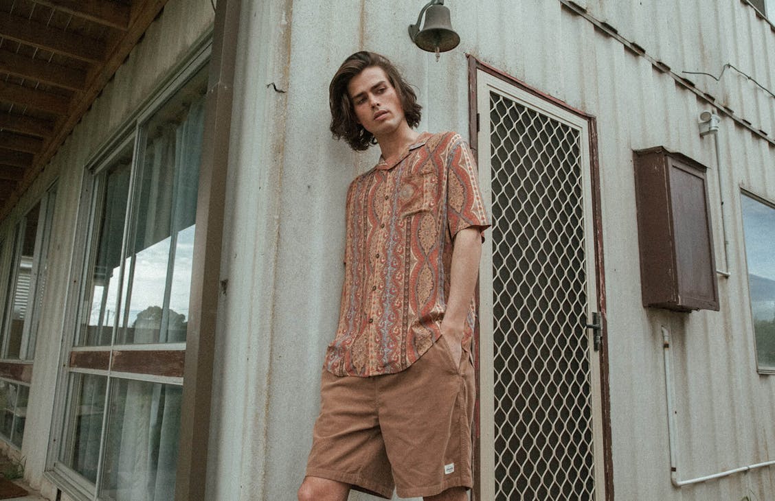 Male model wearing the Fala shorts in Mud and the Bowler shirt in hotel stripe.