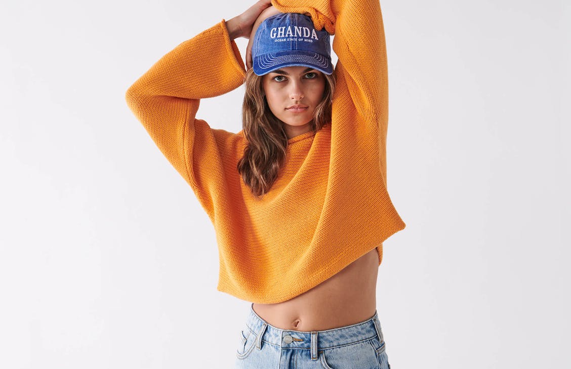 Female model in studio wearing orange knit jumper and classic cap.