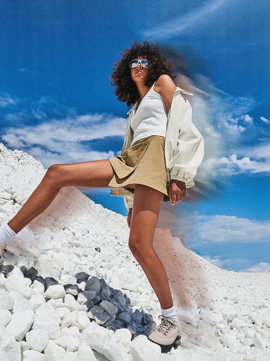 Image of female wearing a brown cargo mini skirt, white singlet and a white bomber jacket. She's stepping up onto a mound of white sand with blue skies behind her.