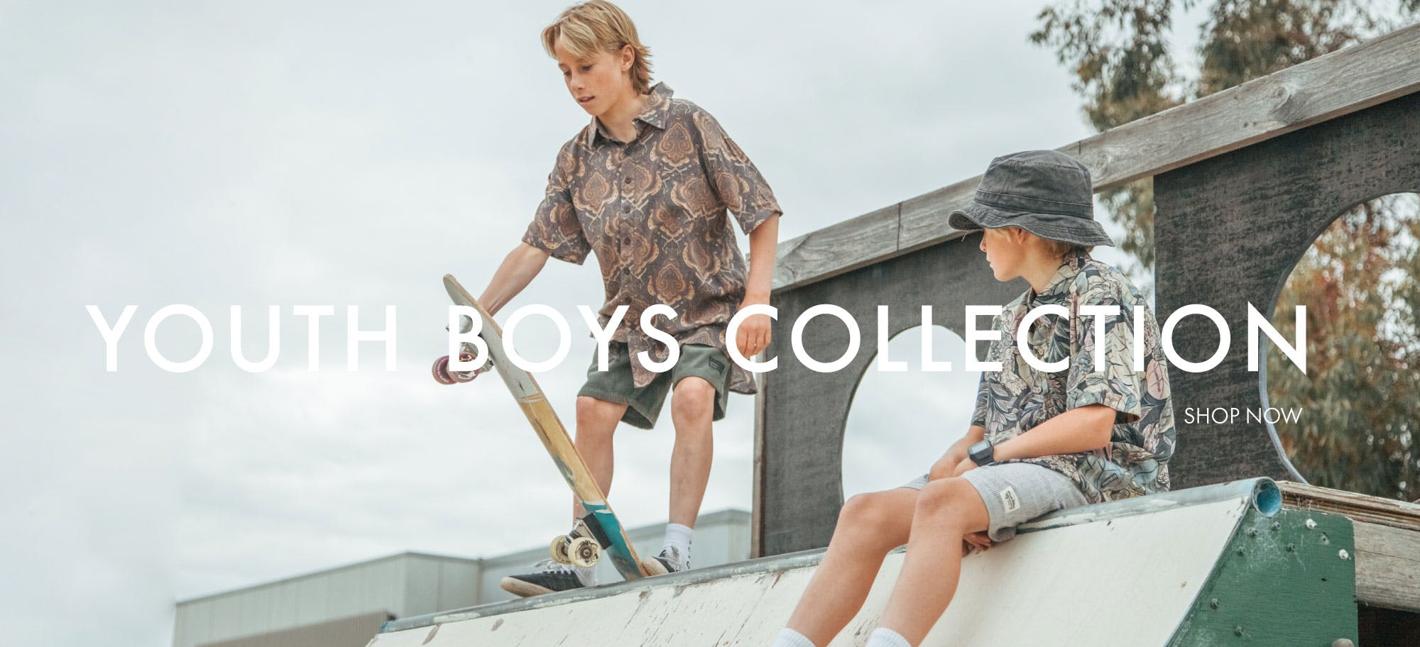 Two teenagers at skate park with Youth Boys Collection in white text overlay. Left boy stands holding skateboard in paisley Shirt, right boy sits with blue Bucket Hat & paisley short sleeve Shirt.