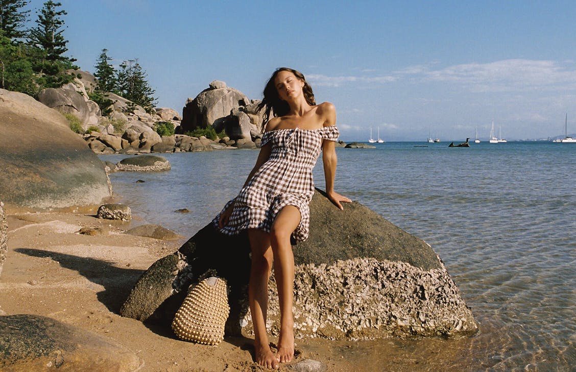 Female model wearing the Peggy dress in bark check leaning on a rock at the beach.