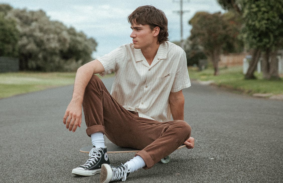 Male model sits on skateboard of asphalt road wearing the Ashton Cord Pants in Barka nd the Donny Shirt in Walter Stripe. 