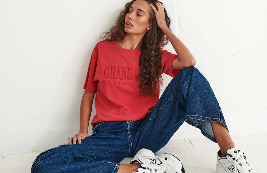model posing in studio. wears a red embroidered tshirt and blue jeans.