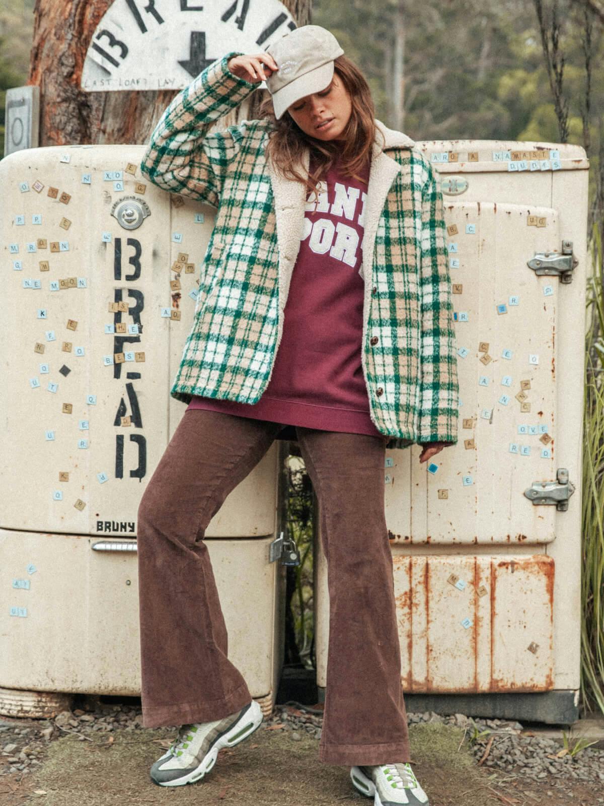 Image of female model standing infront of a bread fridge on the side of the country road. She is wearing the North Crew in Maroon under the Woodland Jacket with the Cord Flares in Brownie. 