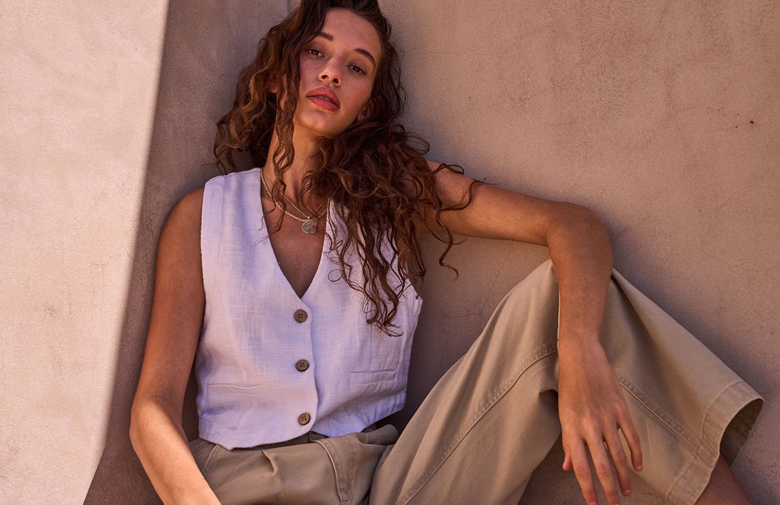 Image of female in a beige concrete courtyard. She is wearing light brown pants and a white linen vest. 