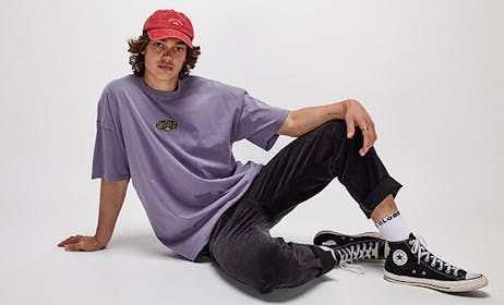 Image of male model in studio, he is wearing black denim jeans, black converse and a purple t-shirt with Ghanda embroidery, he is also wearing a red cap.
