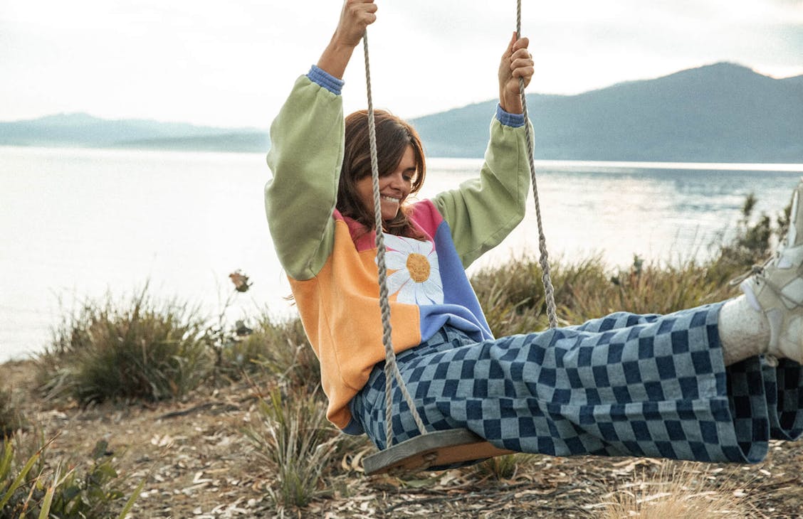 Image of female model wearing the Panel Thrift Crew and Cameron Jeans. She is sitting on an old wooden swing with the ocean landscape behind her. 