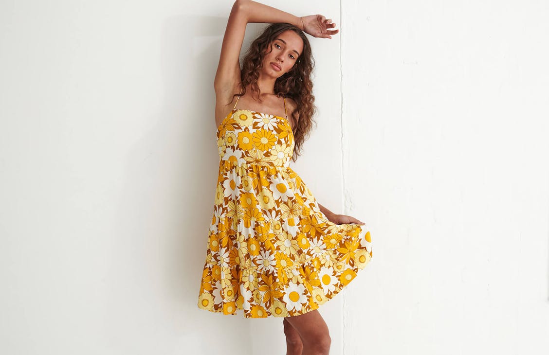 Model wears a yellow and orange floral dress in studio. Poses with arm above her head for photograph