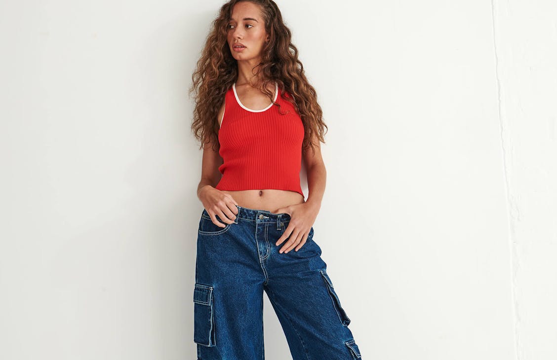 model poses in studio. She is wearing red top with white ribbing and blue denim cargo pants. 