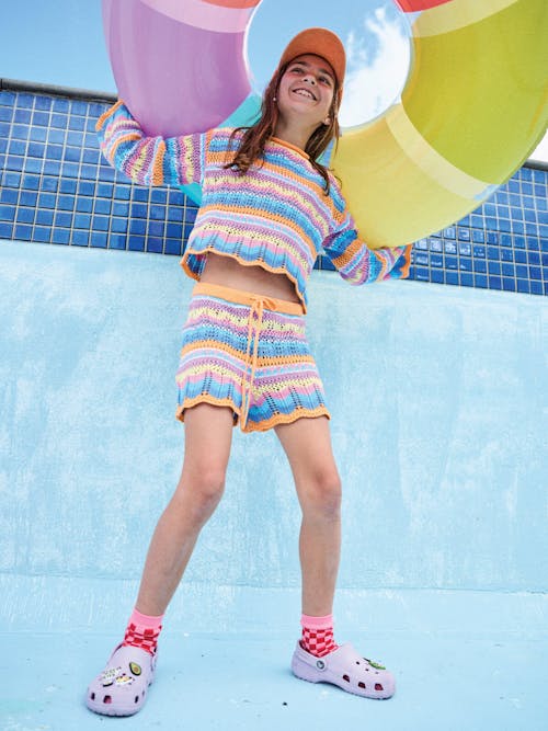 Photograph of girl in matching knitted shorts and top set. Standing in an empty pool, holding rainbow pool ring above her head.