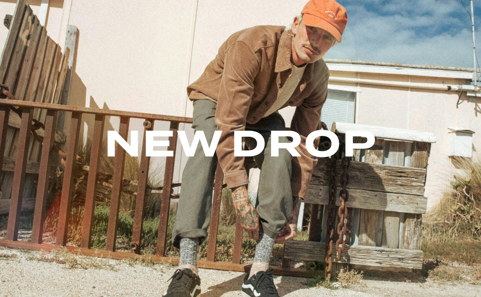 Male model wears the Byron Pants in Fern Green and the Fala Shacket in Desert Khaki with the Classic Cap in Washed Orange. He sits on an old cast iron fence with a cream house in the back. Text overlay: new drop