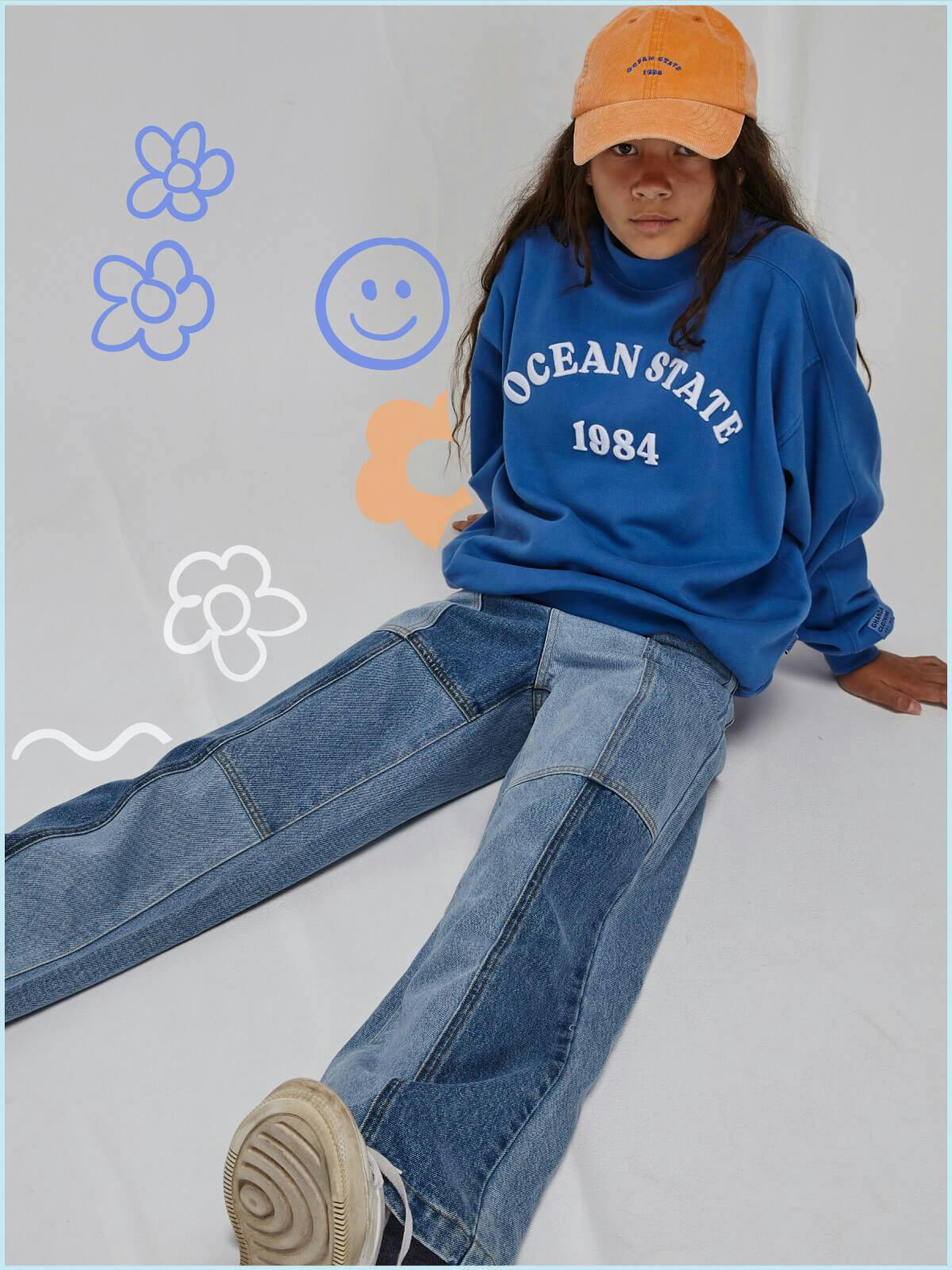 Image of teen girl in studio. She is sitting on the floor and is wearing the Cameron jeans in Patchwork Denim, the North Crew in Baddie Blue and an Orange Cord Cap
