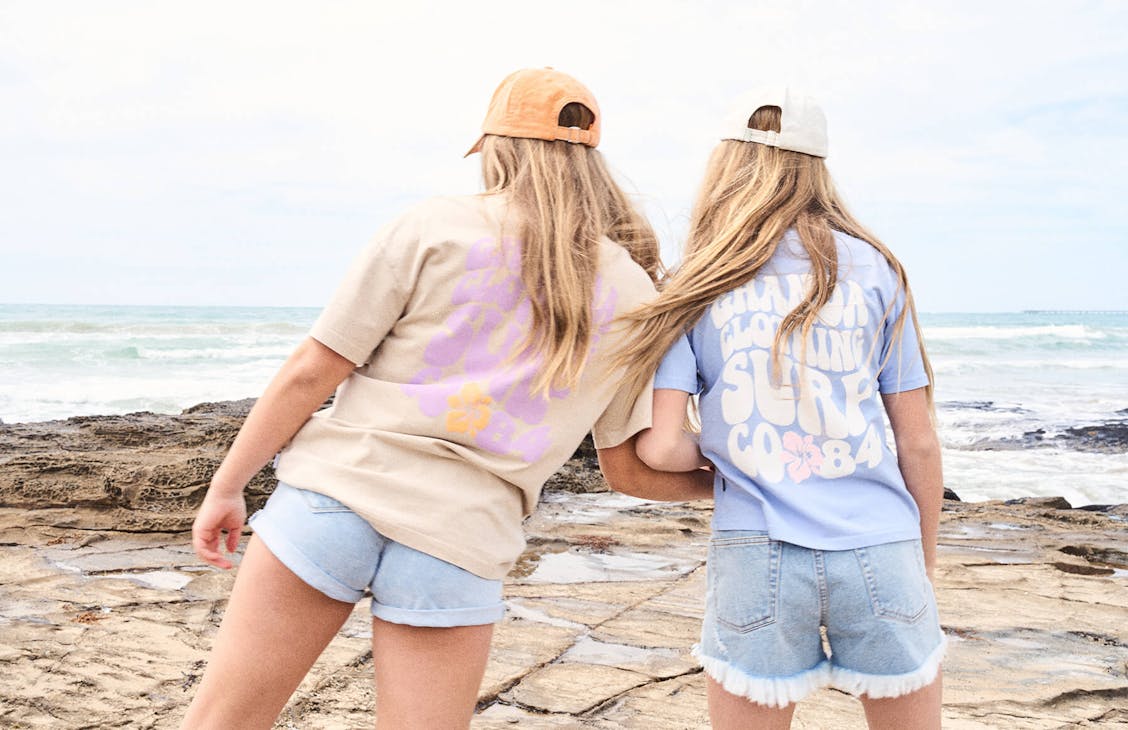 Image of two teen girls holding hands as they walk away form the camera along the rocks at the beach. They are both wearing denim shorts and t-shirts with back print