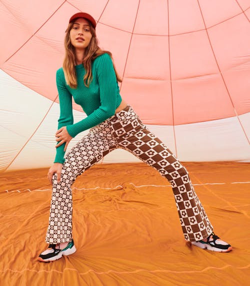 Female model wears the Eva Jeans in Brown Daisy and the Harper Long Sleeve Top in Grasshopper Green. She is standing inside a hot air balloon in wide leg stance leaning to one side.
