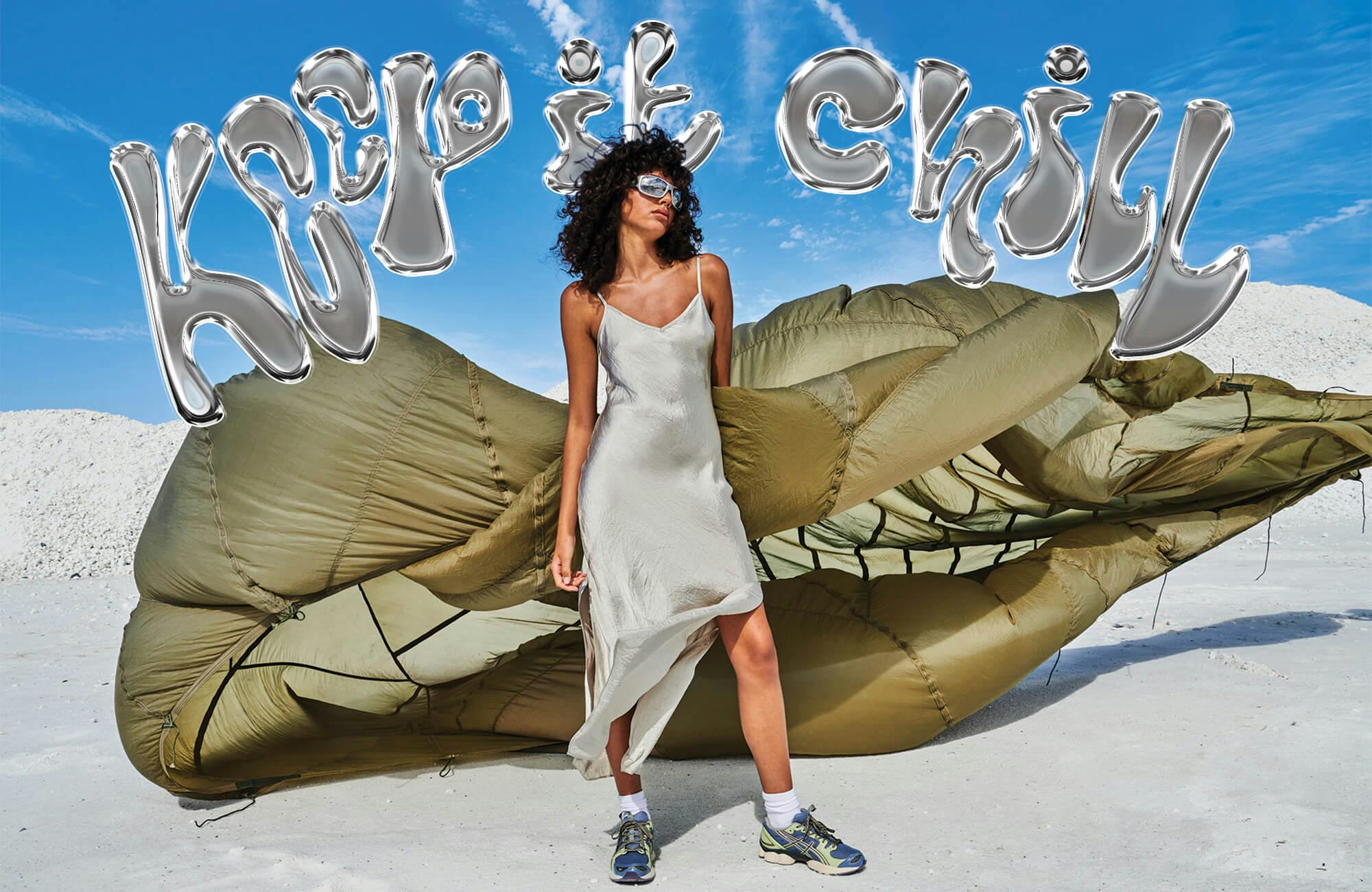 Image of female model standing on white sand, she is holding an army green parachute behind her that blows in the wind, with blue skies. She is wearing sneakers and a silver slip midi dress with silver space sunglasses. Text with 3D silver effect is positioned at the top, reading "keep it chill"