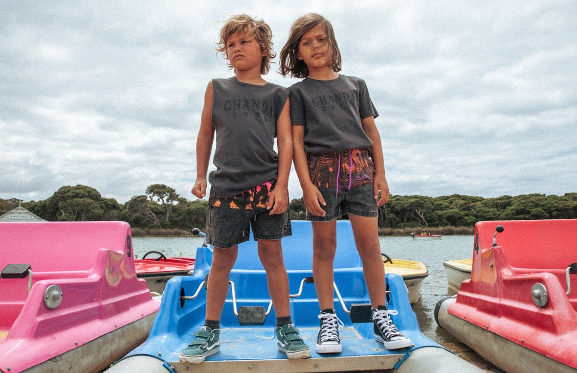 Two young boys stand on a paddle boat. The on on the left wears Jam Shorts in Sunrise Tie Dye with the Thrift Tank. Boy on the right wears Jam Shorts in Electric Swirl with a regular tee