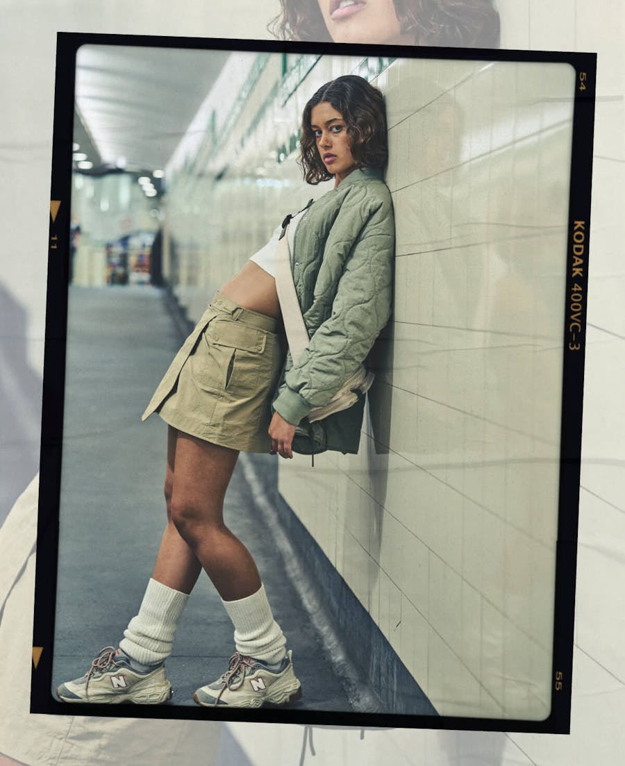 Image of female model leaning on the train station wall. She is wearing a mini skirt, top and a jacket with leg warmers and sneakers. Image has black film frame around it.