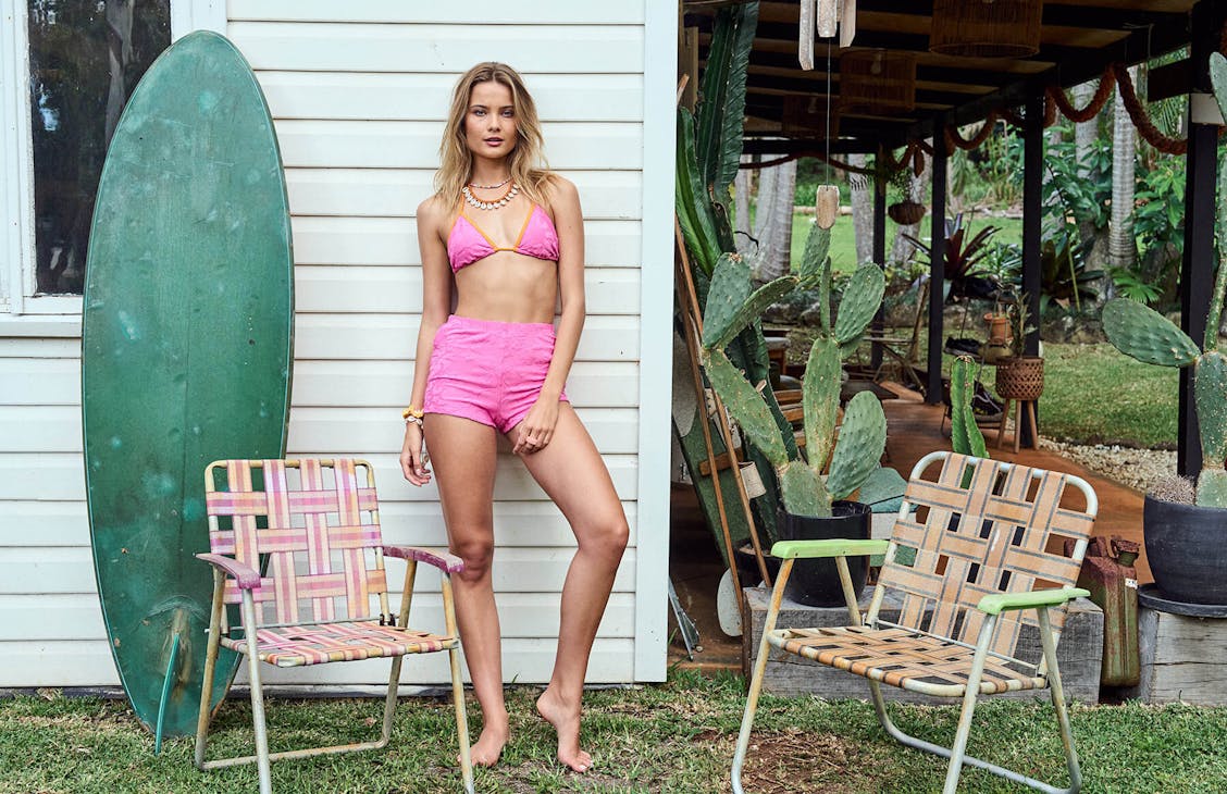 Image of female standing on the side of weatherboard house, around her are lawn chairs and other household items like a surfboard and plants. She is wearing a pink triangle bikini top and matching pink swim shorties. 