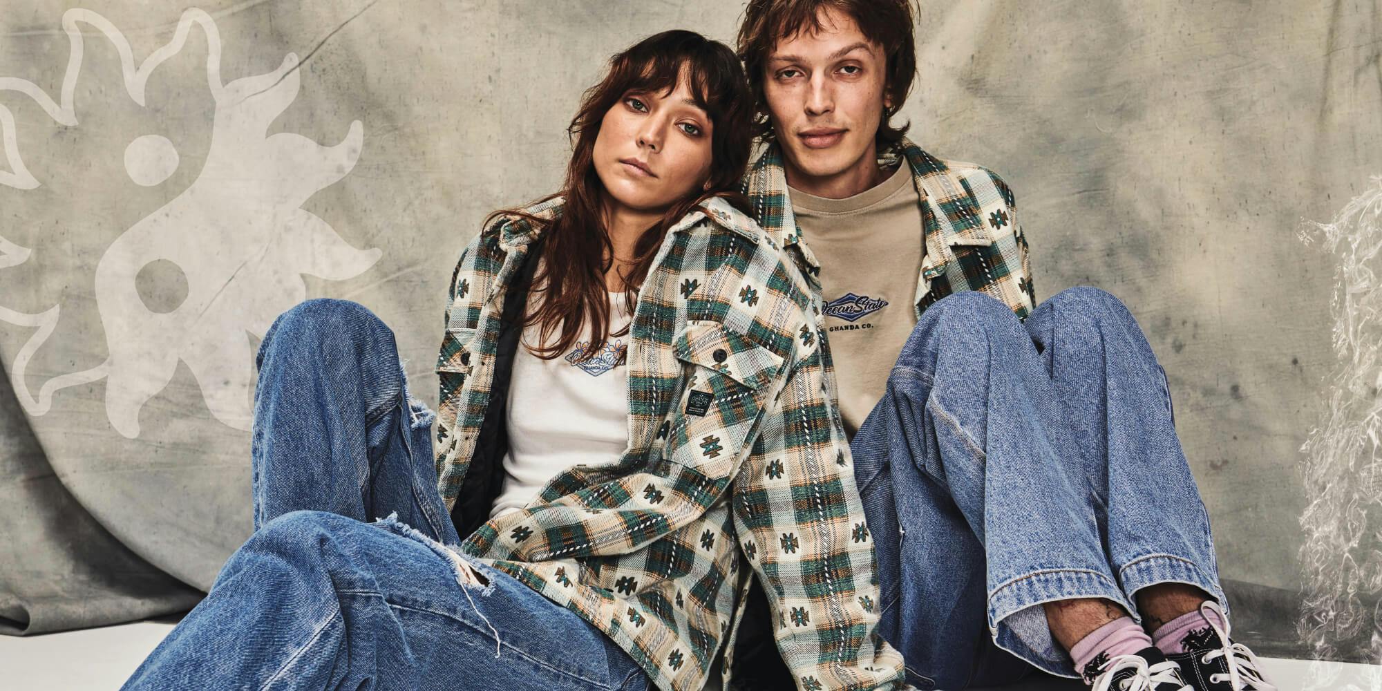 Image of female and male model both wearing denim jeans and a checked jacket, they're sitting in front of a light wash denim background.