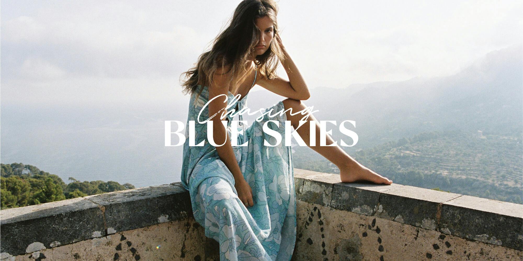 Image of female sitting on a blankly with Spain in the background, she is wearing a blue floral maxi dress. ˇext overlays reading 'chasing blue skies;