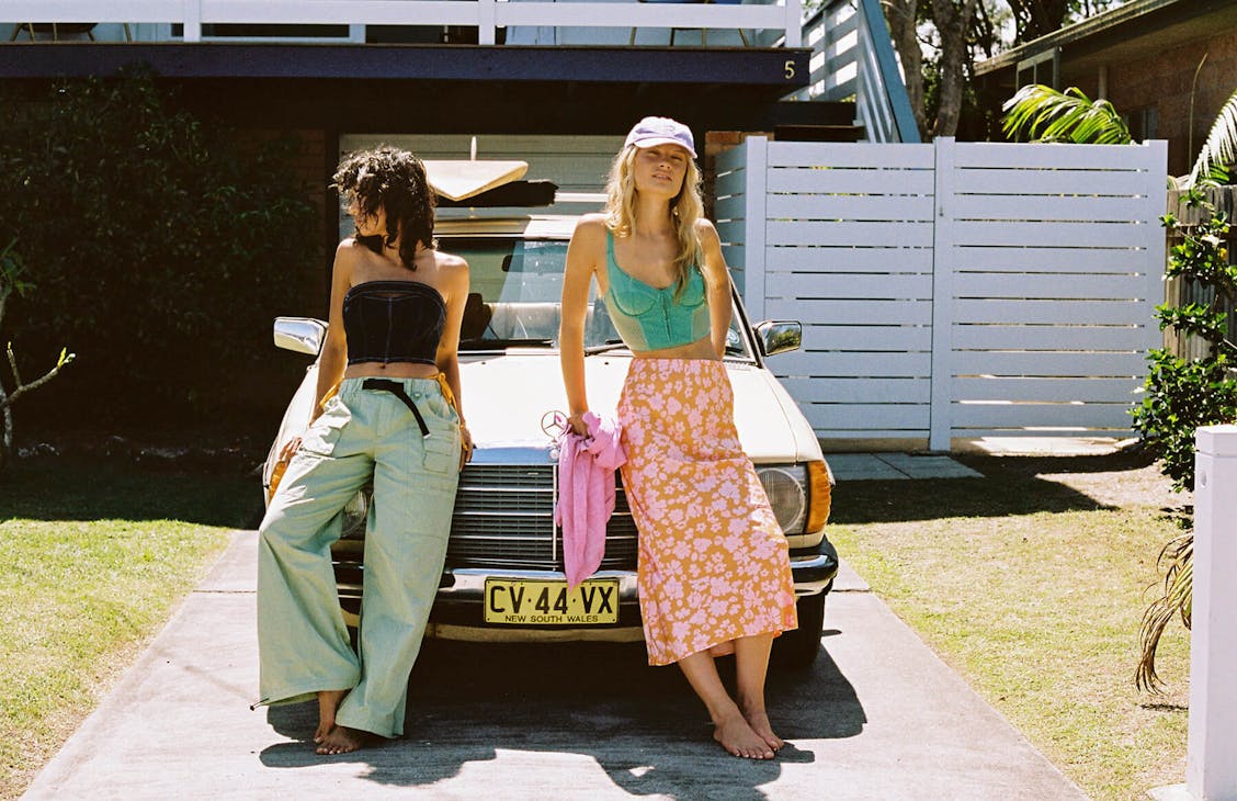 Image of two females leaning on the the bonnet of an old Mercedes in the front yard. Model on the left wearings green parachute pants and a strapless denim corset top/ Model not the right wears a orange and pink floral skirt with a green lace corset top and purple cap