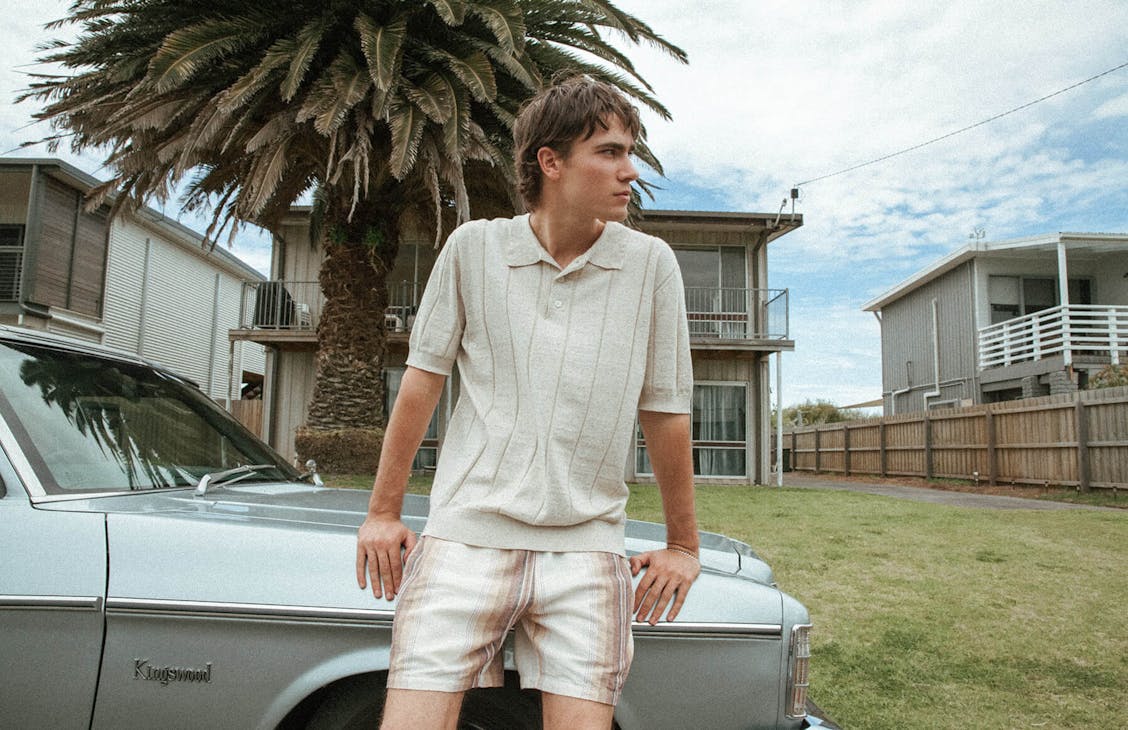 Male model wearing the Pop polo in tuna natural with the Murphey shorts in Ashton stripe, leaning against the Bonet of an old car. 