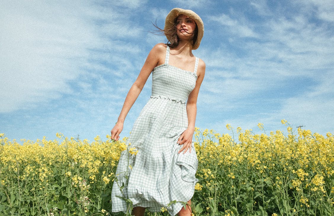 Female model stands in canola field wearing the Brighton Dress in eggshell check and the sunshine rafia hat in natural
