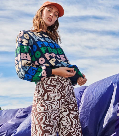 Female model stands in front of blue cloudy skies and blue parachute, she is wearing the Party Pants in Mud Pool Swirl and the Fiesta Knit and Classic Cap.