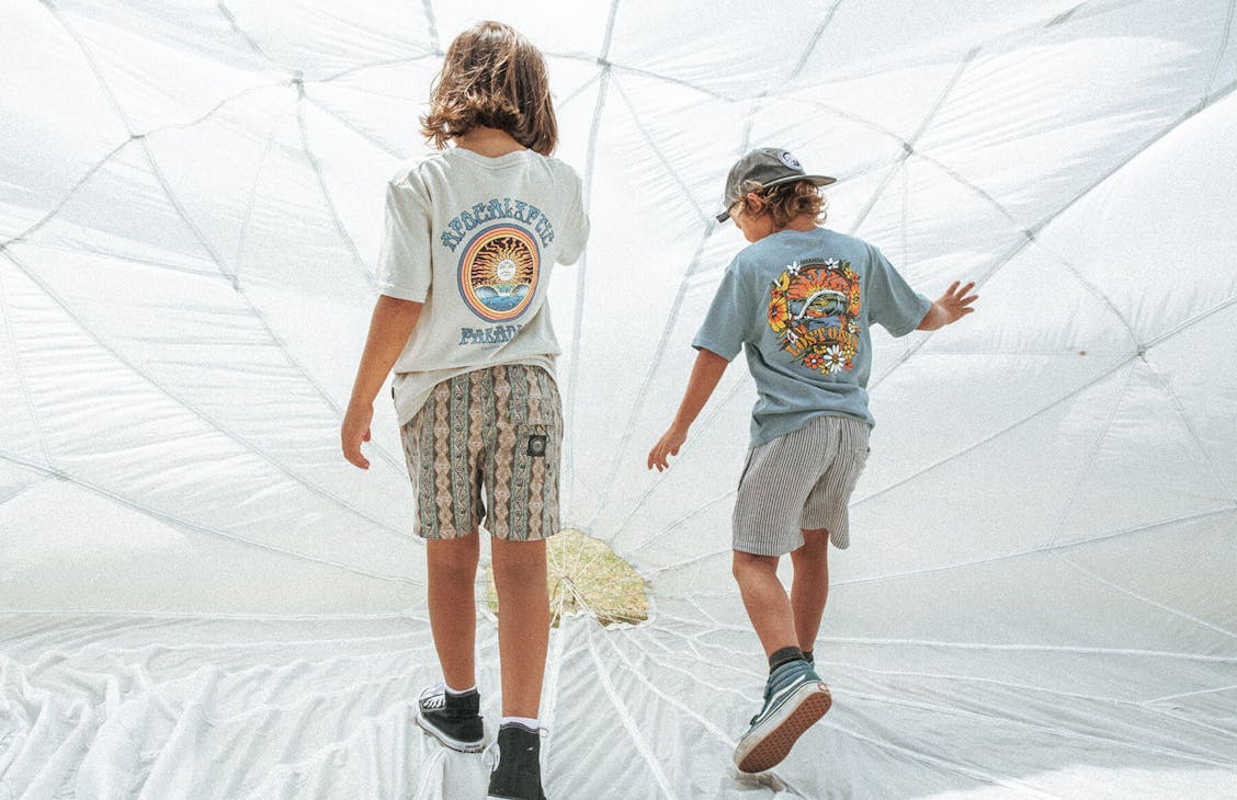 Two young boys play under an open parachute. The boy on the right wears the Jam Shorts with the Fireball Vintage Tee and the boy on the right wears Ruben Shorts in Prison Stripe with the Oasis Surf Tee