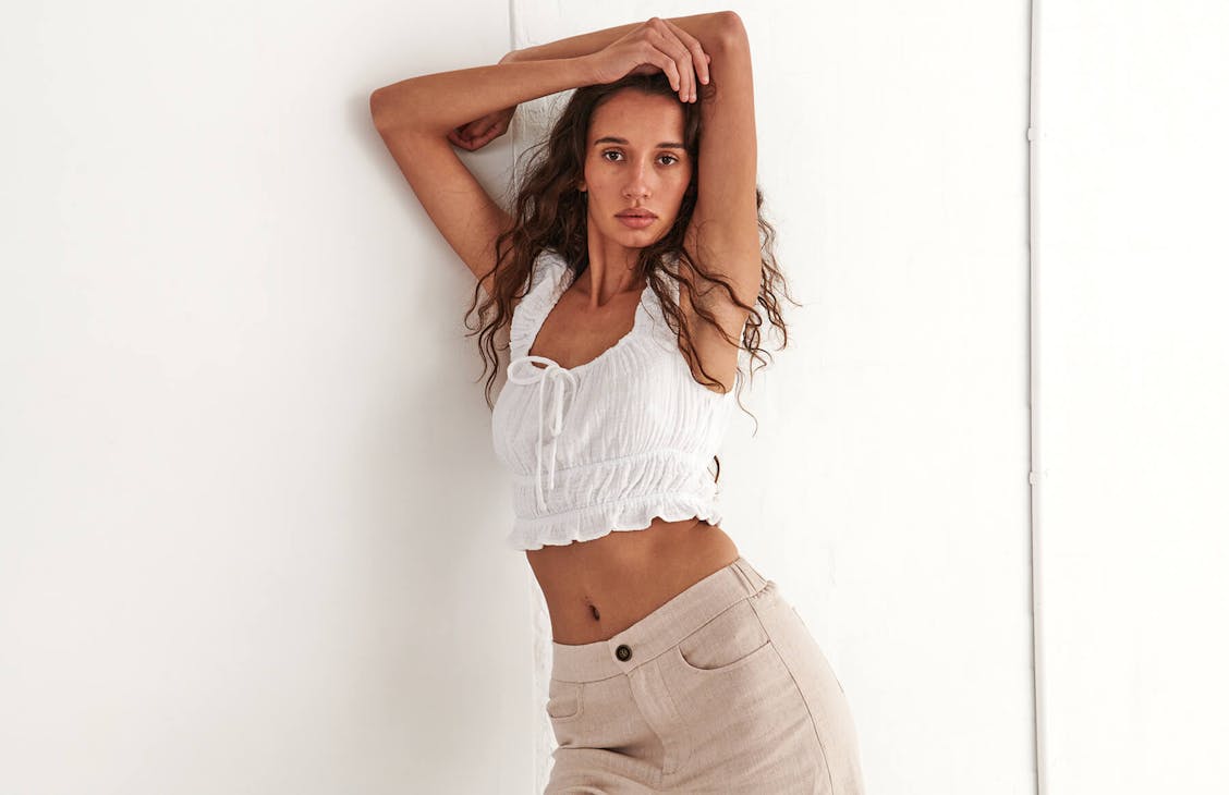 Image of female model in studio, she is wearing a white cap sleeve crop top and beige pants. 