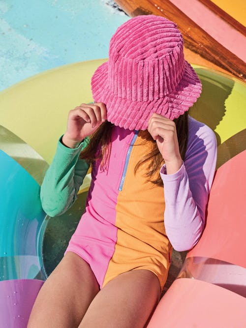 Photograph of girl in colourful long-sleeve swimsuit, and pink hat covering her face. She sits in rainbow pool ring.
