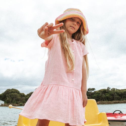 Young girl wears the Rockpool dress in pink standing on a yellow paddle boat making a peace sign with her fingers. 